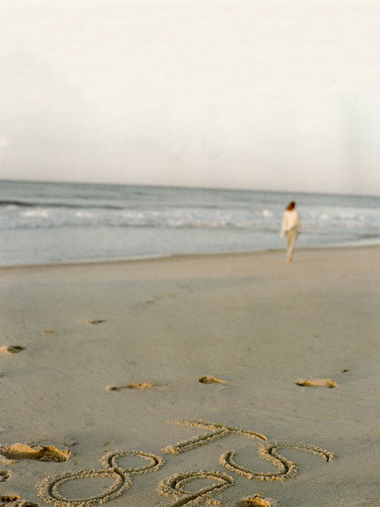 1989 TV Taylor Swift. She’s pictured during the 1989 TV album photoshoot. She is walking far away on a beach, close to the waterline. She’s wearing light wash jeans and a white button down. In the sand are her footprints and “TS 89” written in the sand. 