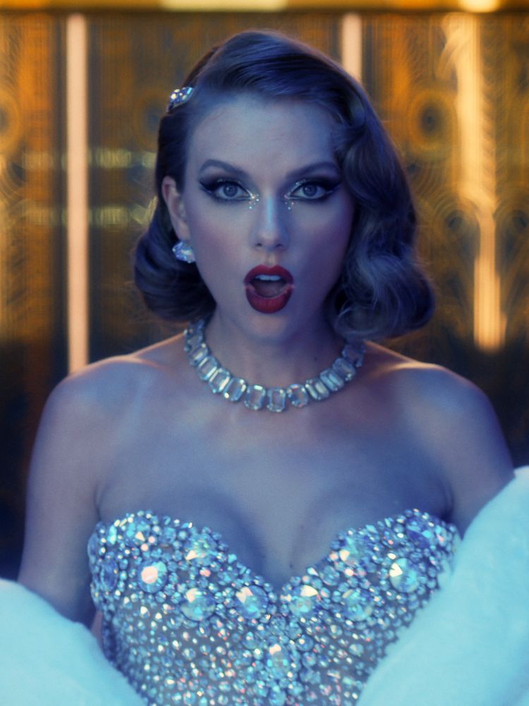 Midnights era Taylor Swift. She is pictured in the Bejeweled music video. She is wearing a bedazzled bodysuit with a fur shall. She has matching jewel necklace and earrings and is in stunning, dark red lipstick. Her eyes are dramatic with cat eye liner and sparkles. Her hair is short and pinned with jewels. 