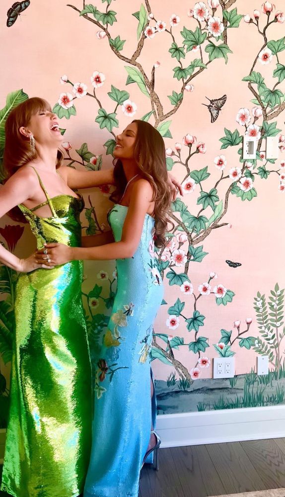 Taylor Swift and her friend, Keleigh Teller (Miles Teller’s wife; both starred in the I Bet You Think About Me music video) posing and laughing. They stand in front of a beautiful wall depicting branches, flowers, butterflies, and other greenery. Taylor is in a fabulous green sequin dress with neutral makeup and her hair down in waves. Keleigh is wearing a blue dress with embroidery work, with neutral makeup and her hair down in waves. 