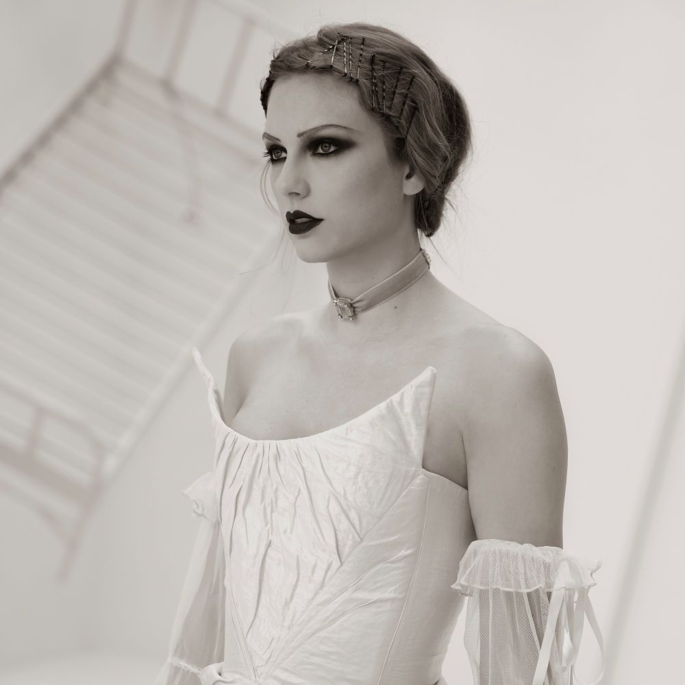 Taylor Swift in the Fortnight music video. She is in the white dress standing in front of the bed railing in a white room. Her hair is up and she has dark, dramatic makeup on. 