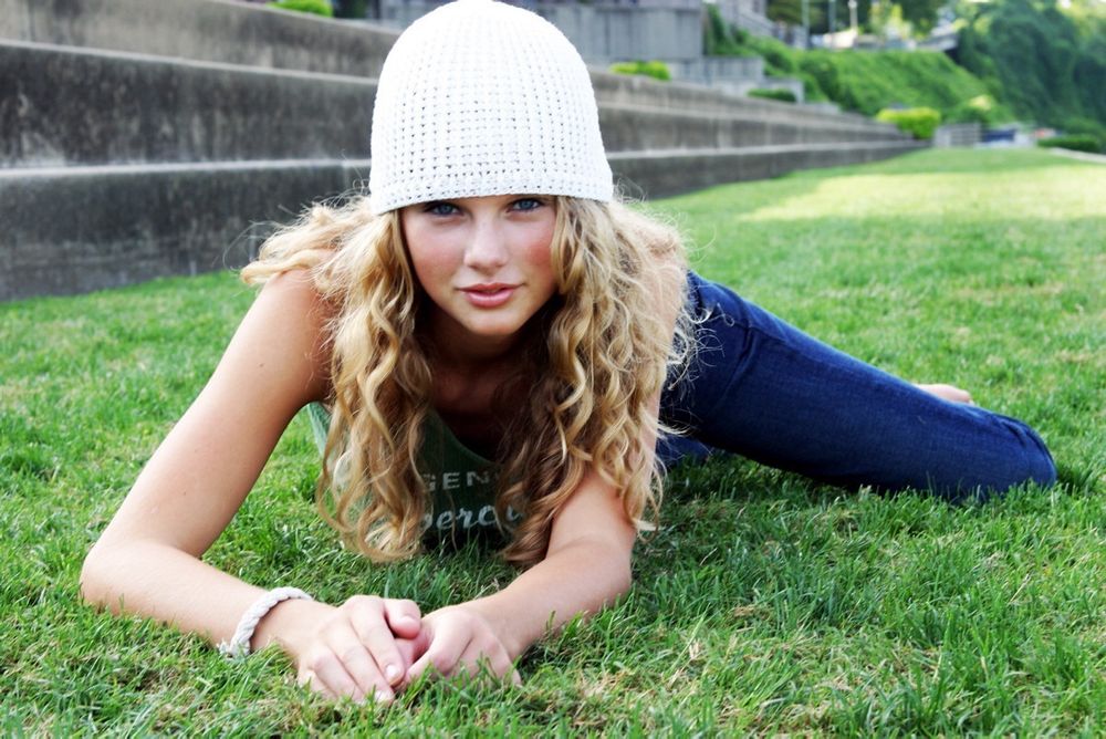 Young Taylor Swift photographed laying on her stomach in an army crawl position in the grass. She has dark blue jeans, a green ambercrombie tank top, and a white knit hat on. She has her natural curls down and is wearing minimal makeup and a twist bracelet. 