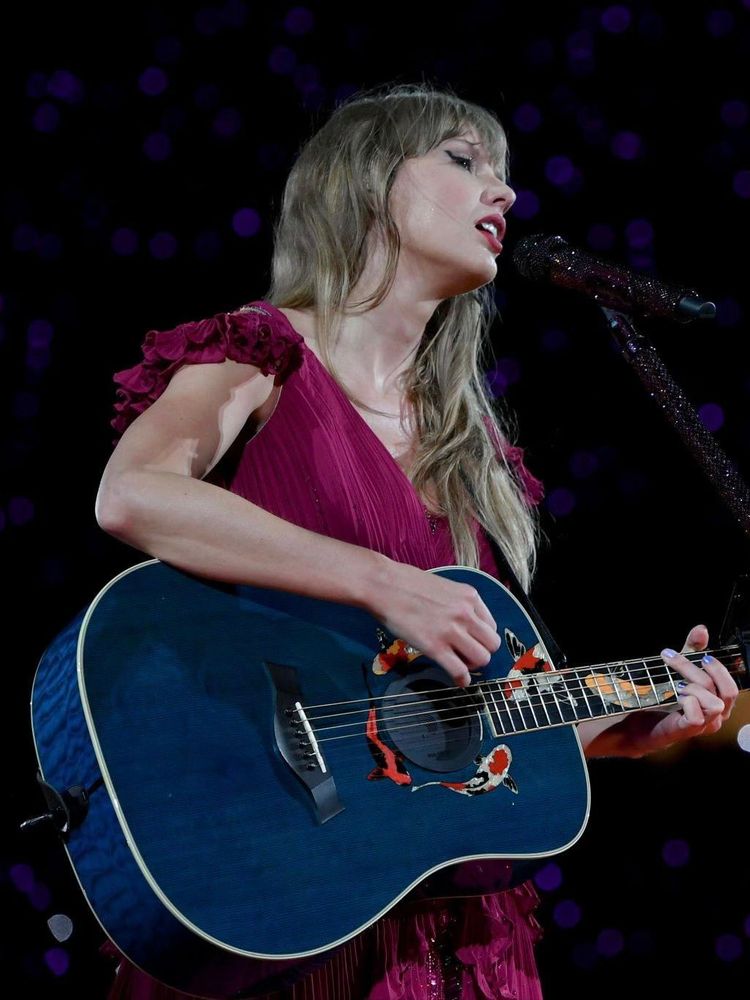 Speak Now TV era Taylor Swift. She’s on stage at The Eras Tour, performing during the surprise song set. She’s is holding the Speak Now era blue koi fish guitar while wearing the original red surprise song dress. 