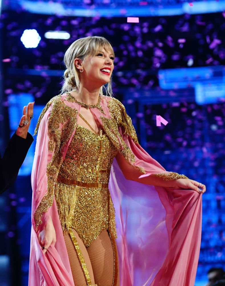 Taylor Swift accepting an award at the AMAs. She has a pink and gold cape on over her golden bodysuit. She’s smiling to the crowd. 