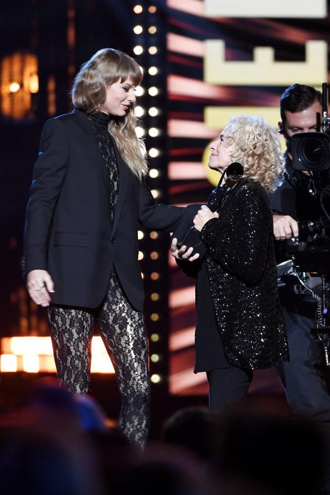 Red TV era Taylor Swift. She is on stage honoring Carole King at the Rock and Roll Hall of Fame. Taylor is wearing a lace bodysuit with a black blazer over the top. Carole is looking up at her while holding her award; she is wearing a black dress and tights with a sequin black cardigan over the top. A camera man is pictured beside them. 