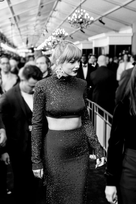 Midnights era Taylor Swift at the 2022 Grammys. This black and white photo shows her on the red carpet. She is wearing the iconic dark blue sequin two piece dress, dramatic large earrings, dark makeup, and her hair up in a sleek bun. 