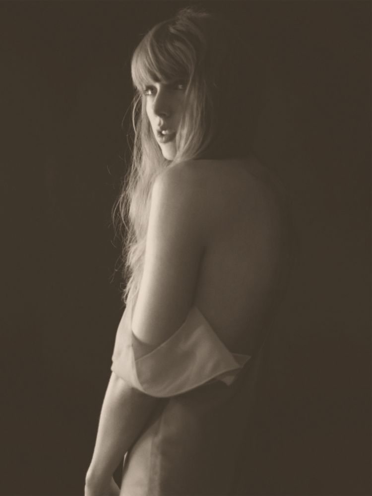 TTPD era Taylor Swift. She is pictured for the TTPD album photoshoot in a sepia tone. She’s standing in a loose nightgown with the shoulder falling off her arm. Her hair is long and wavy as she looks over her shoulder. 