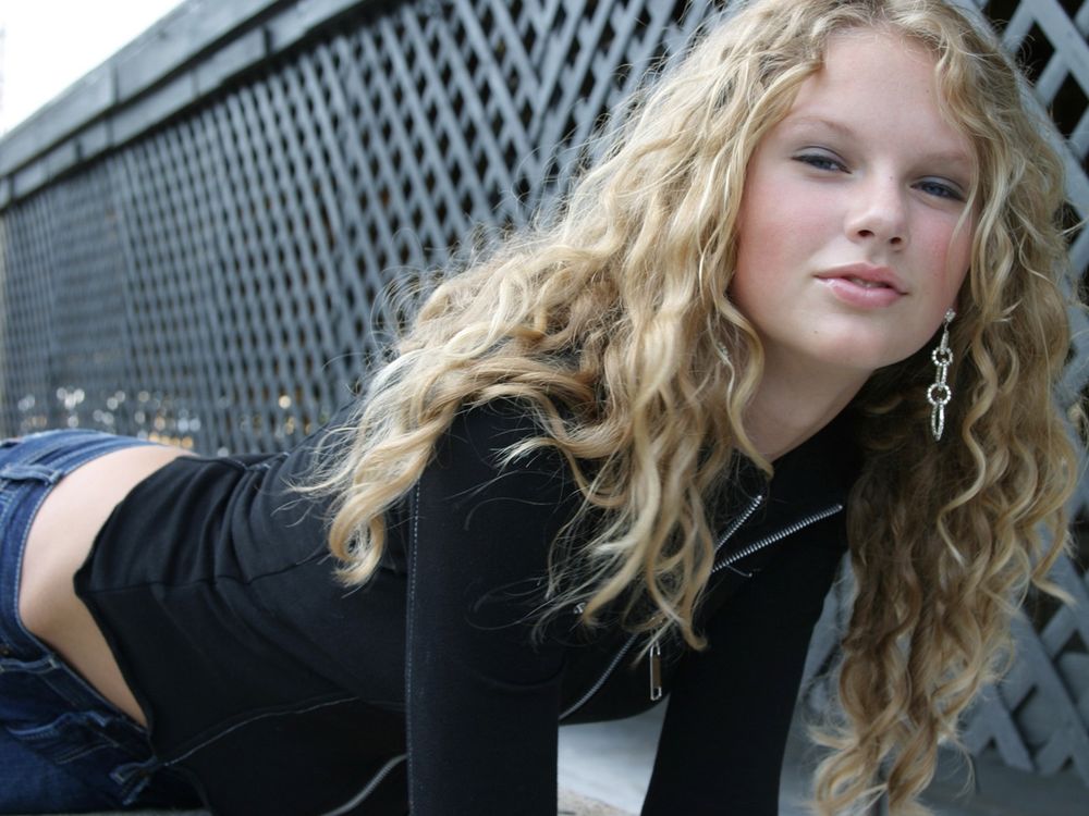 Young Taylor Swift photographed leaning to the side in front of a fence. She has dark blue jeans and a black zip up on. She has her natural curls down and is wearing minimal makeup and silver earrings. 