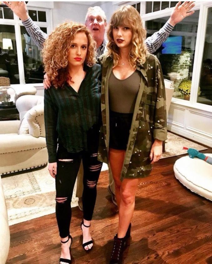 Taylor and Abigail posing for a photo. Scott, Taylor’s dad is photobombing them. Taylor is in a camo jacket, army green top, black shorts, and black shoes. Abigail is in a striped black and green button down, ripped black jeans, and black heels. 