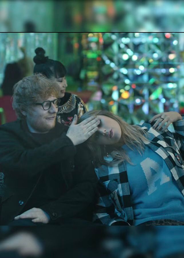 Taylor Swift and Ed Sheeran in the End Game music video. Ed is in a black outfit and glasses, patting Taylor’s forehead. Taylor is in a plaid button down with a blue sweater underneath. Her sweater has a white A on it, likely a nod to Ed’s song, A Team (which is also used as a lyric in the chorus of End Game). Taylor is laying her head on Ed’s shoulder. 