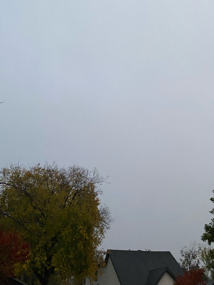 Picture of overcast clouds, with the roof of a singular house at the bottom of the picture. The house is surrounded by trees next with red and green leaves.