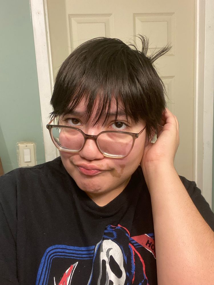 The image has a person with short hair posing. They smile seductively, pursing their lips and partly smiling. With their hand behind their ear as if they were pushing back long luscious hair. Staring in a way where their glasses and face are lowered and eye line is over the lenses. With their hair sticking up at the crown of their head. The hair is standing in a way where it forms to straight clumps in the shape of a letter v.