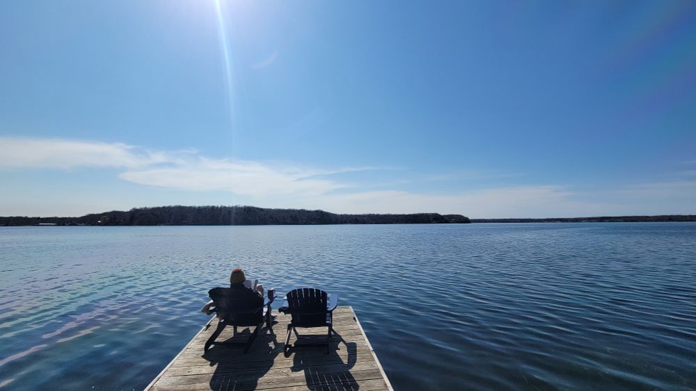 A person reading in one of two adirondack chairs on a dock on a lake.
