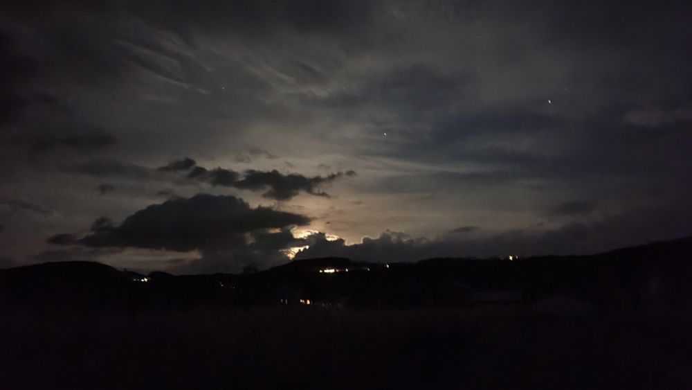 A cloudy night sky with hills on the horizon. There are a handful of artificial lights and a few stars visible. A nearly full moon is coming over the hills, mostly hidden by dark clouds