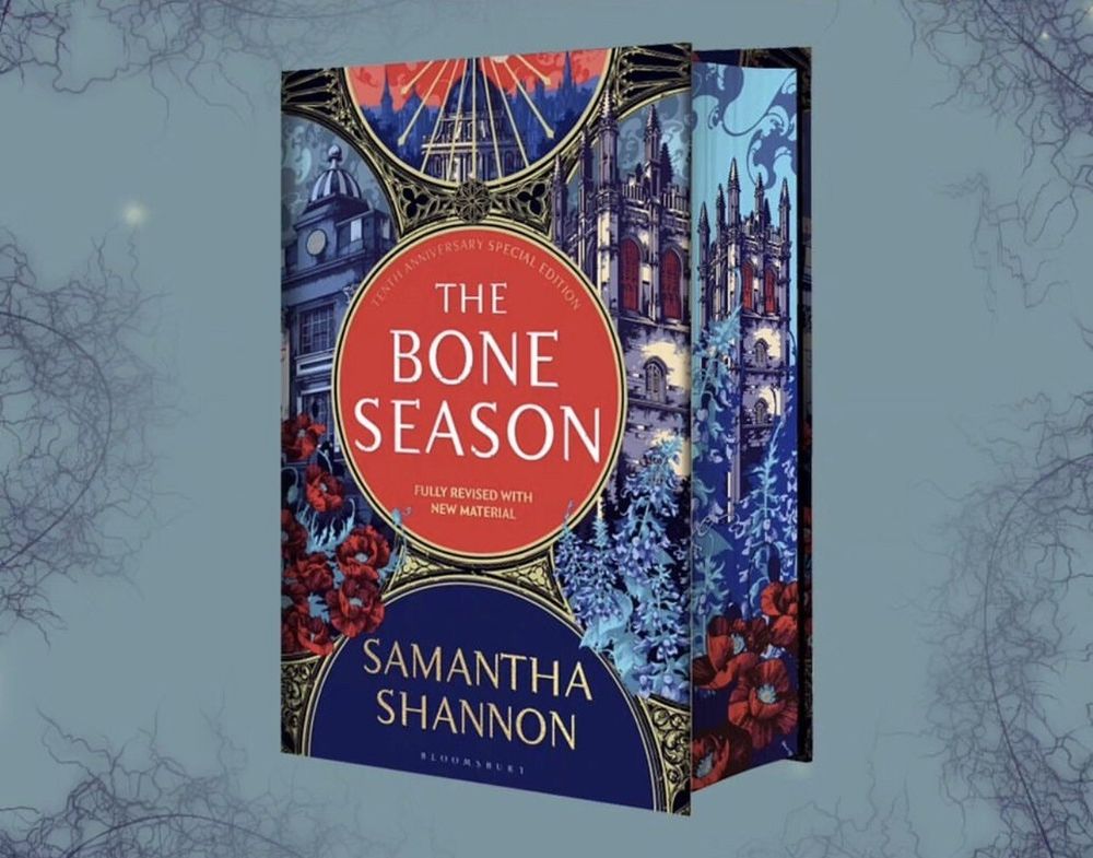 Superbe couverture de l'édition spéciale 10 ans de "The Bone Season" par Samantha Shannon. Design ultra détaillé avec des bâtiments anciens, des fleurs rouges et des touches dorées sur fond bleu. Le titre ressort bien au centre dans un cercle rouge.