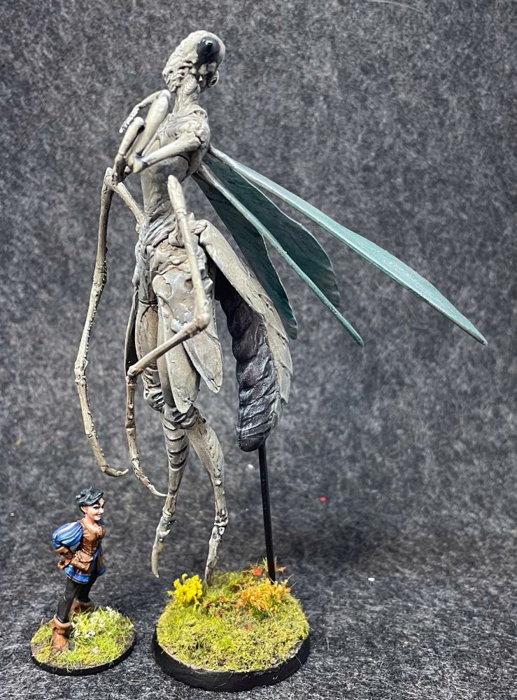 A large praying mantis miniature, next to a 32mm scale mini of a human.  She's at least 4 times taller than the human 😅
The human seems oddly unconcerned about this ..
