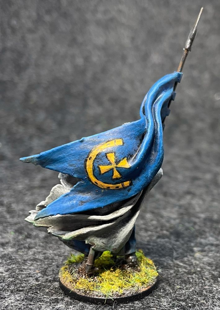 Painted miniature of a knight wearing chainmail, tabard and a woolen cape, carrying a peaked helmet with a nose bar in one hand, a banner in the other. 
His tabard and banner are blue, and bear the gold (yellow) horseshoe and cross heraldry of the Polish Jastrzębiec family. 
He has streaks of grey in his dark beard