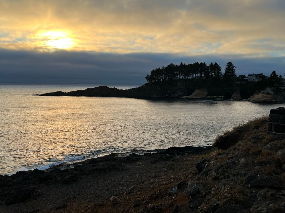 Gorgeous ocean scene at Depoe Bay, Oregon with a tree-lined cliff, setting sun, and a house that some sugar daddy or Dickensian benefactor needs to buy me