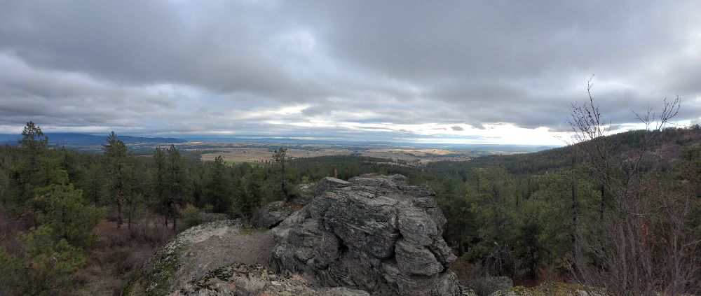 A panorama, featured centrally a large rock on top of a hill with forest on either side. In the distance, you can see plains. It is a cloudy day.