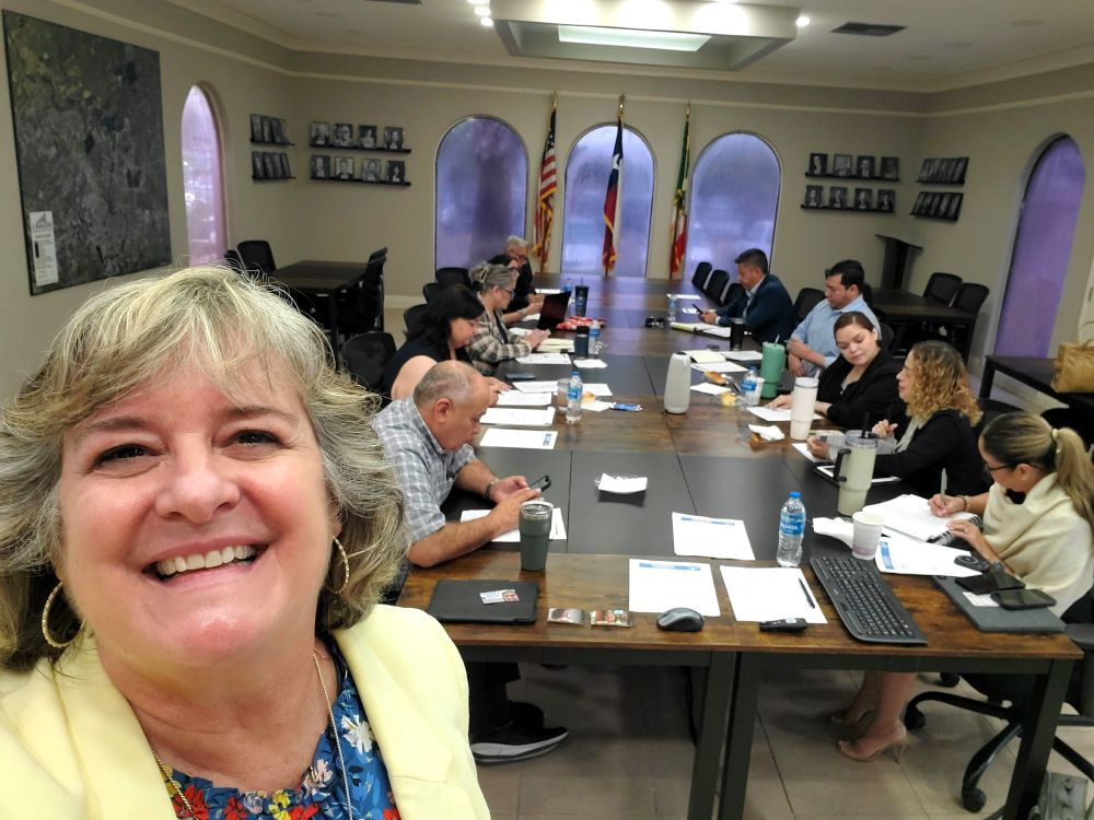 Smiling woman in yellow jacket in front of people working at a table during a social media / digital marketing workshop for an EDC (Economic Development Corporation) 