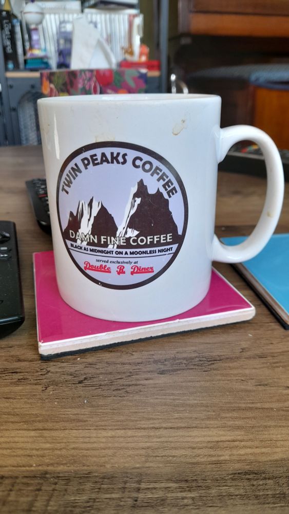 A coffee cup with Twin Peaks Coffee written on it with a picture of two mountains. Underneath it says Damn Fine Coffee and Double R Diner in reference to the TV show.