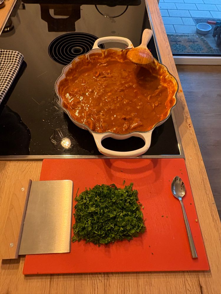 A pan with mushrooms in brown sauce cooking on a stove. A heap of parsley is waiting to be added.