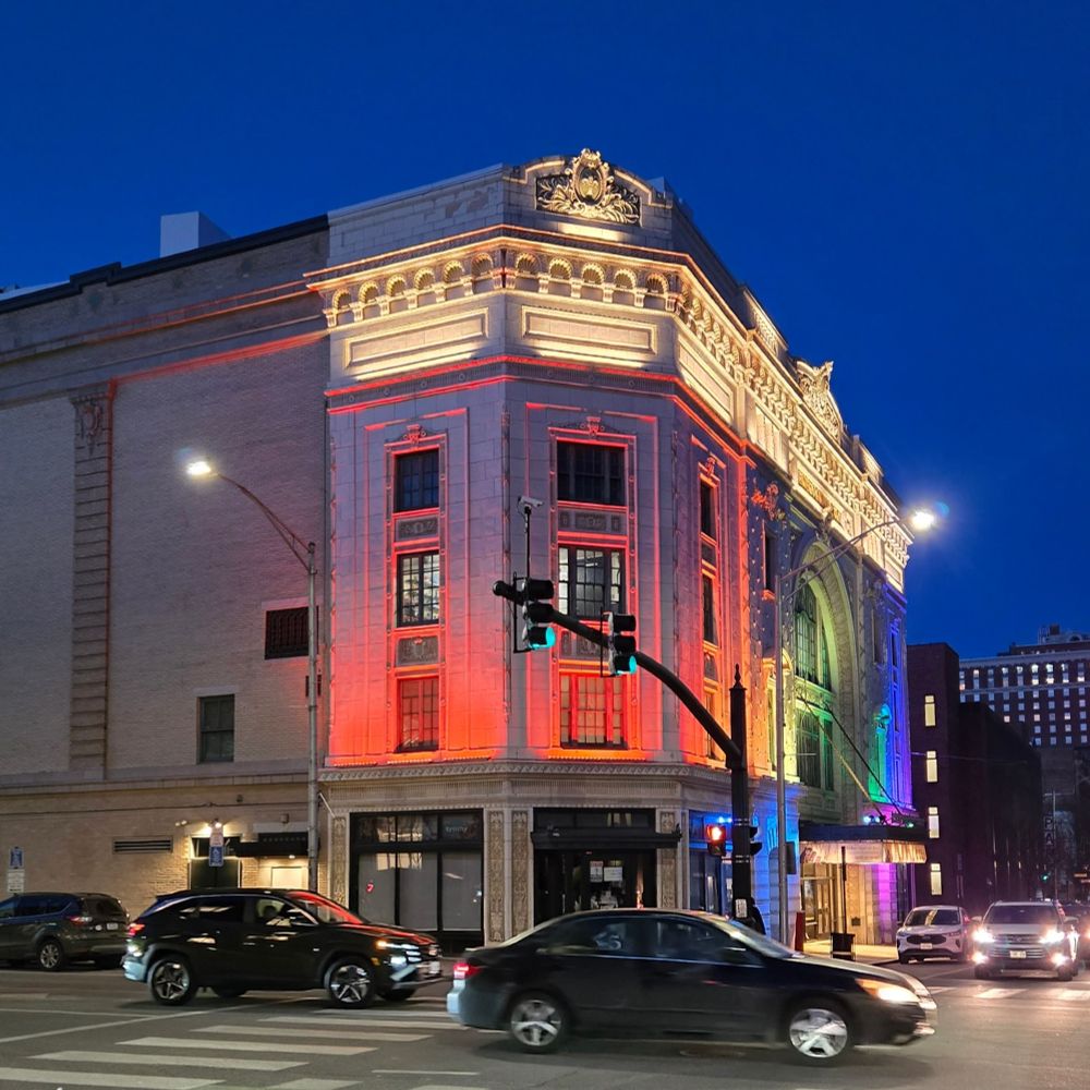 Trinity Repertory Company, a Beaux Artes building in downtown Providence, Rhode Island, is list with a series of colored lights, making a rainbow across the facade. Cars pass by in the foreground.