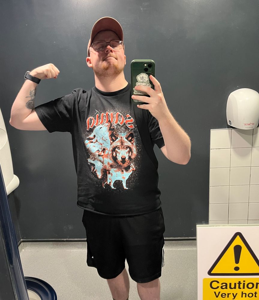 picture of george in a bathroom mirror wearing a hat, a Samia band tshirt and black gym shorts. The sticker in the bottom right of the mirror is cropped and says 'CAUTION VERY HOT'
