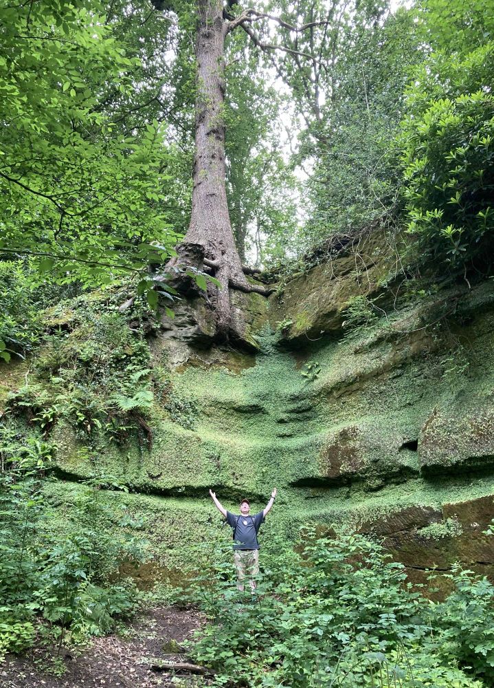 a photo of tiny george standing at the bottom of a large rock outcrop thats covered in mossy vegetation in the woods. Theres a tree growing out of the top, and it's roots are showing. I'm standing with my arms raised high in praise 