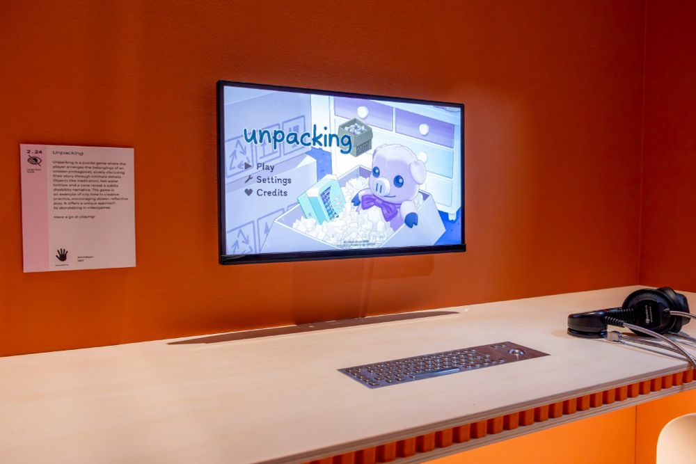 A close up of a computer showing the Unpacking main screen, a keyboard, headphones, and a plaque describing the game and its disability representation through items unpacked, including a hot water bottle, medication, wrist brace, cane and more