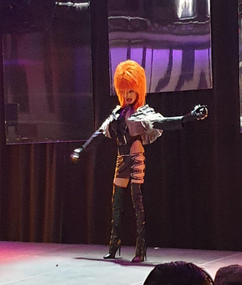 Drag artist Tristana Gray Martyr, wearing a massive ginger wig, and short leather skirt, long boots. Black clothes hidding barely anything