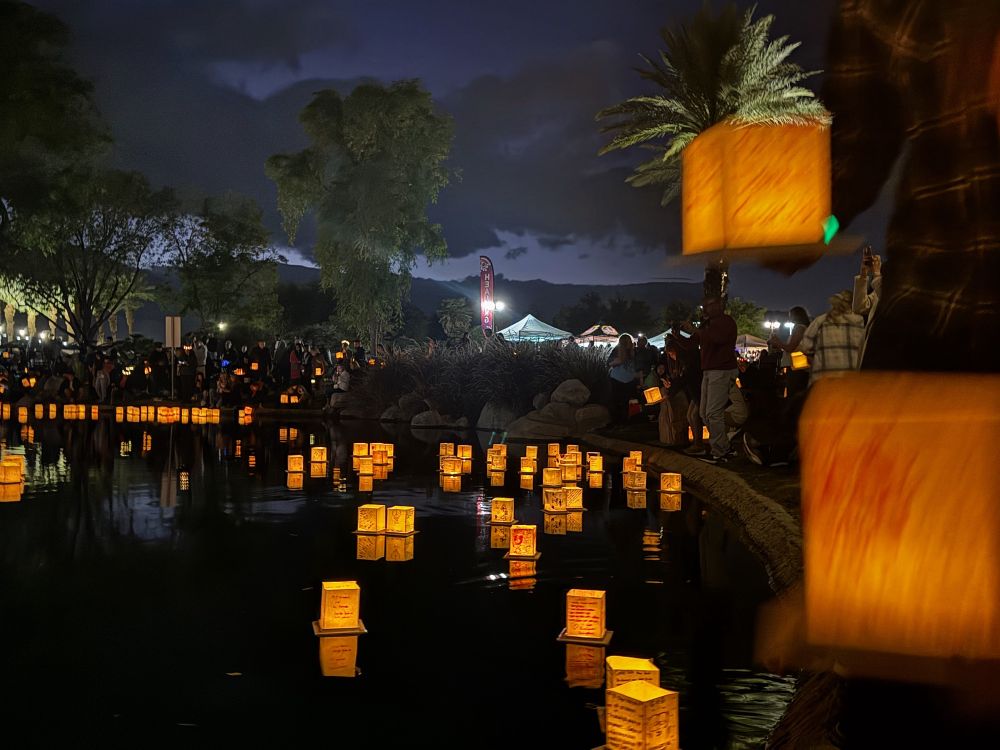 Image of hundreds of square paper lanterns, each lit from within, floating on a calm river after sunset, glowing. Palm trees surround the water, and people with more glowing lanterns. Mountains are silhouetted in the background.