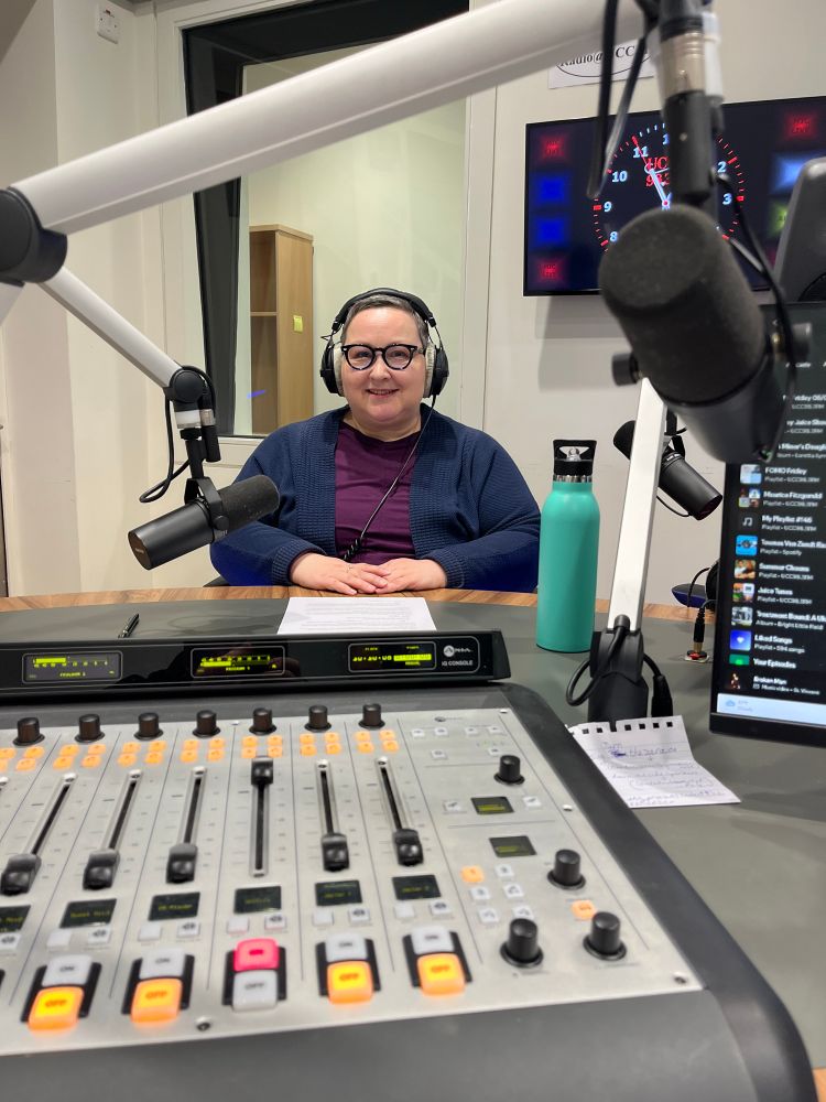 Luigina Colfi, wearing large headphones and glasses, sits at a radio studio desk in front of a microphone. She is smiling and facing the camera. A mixing console with brightly lit buttons and sliders is in the foreground. A water bottle and notes are on the desk, and a studio screen with a clock and “UCC 98.3FM” is visible in the background.