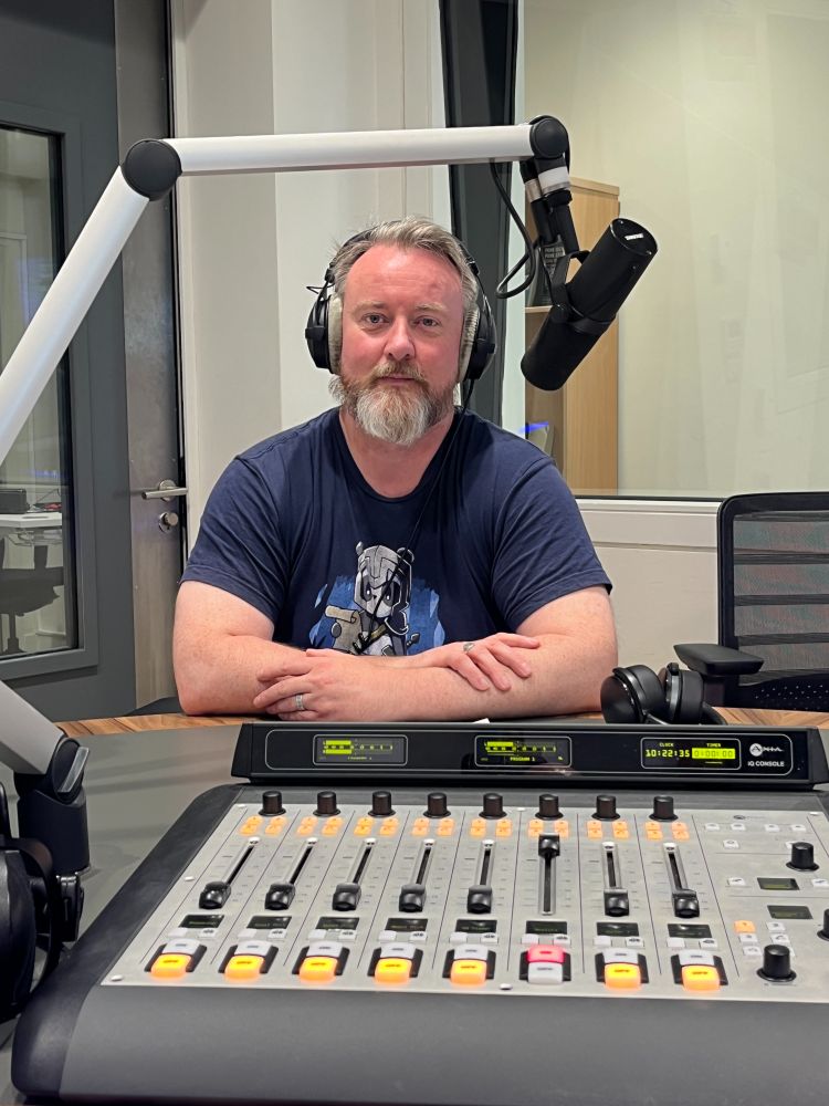 Maurice Ryder is sitting in a radio studio, wearing headphones and speaking into a microphone mounted on an adjustable arm. In front of him is a sound mixing console with various buttons, knobs, and sliders. They are wearing a dark blue t-shirt with an image on it. The background includes soundproof walls and a window into another room