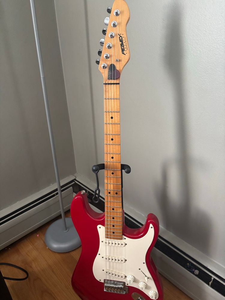 A red Peavey Predator Strat guitar with a white pick guard sitting in a guitar stand by a wall near my lamp. 