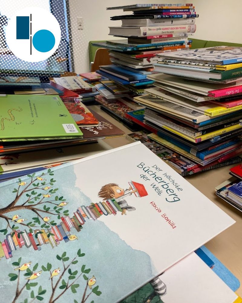 Das Bilderbuch "Der höchste Bücherberg der Welt" liegt im Vordergrund auf einem Stapel. Im Hintergrund sind weitere hohe Bücherstapel.