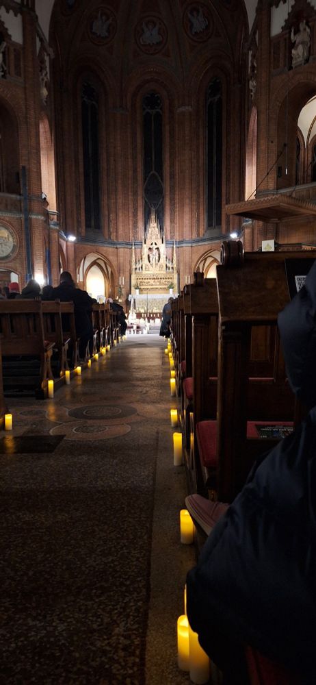 Der Mittelgang einer Kirche. Links und rechts stehen Kerzen auf dem Boden. Der Altar ist beleuchtet. 