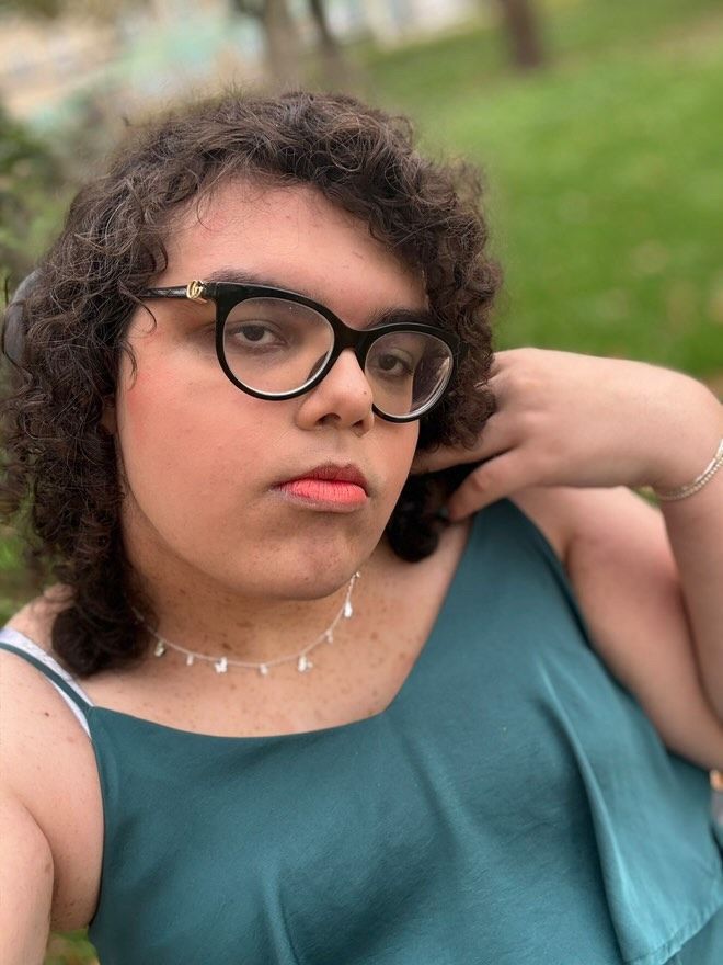 This photo is of me (Hicup) it shows me in front of some green grass with my hand near my head. I am wearing a green dress and a necklace. On my face there are glasses which are oval shaped.
