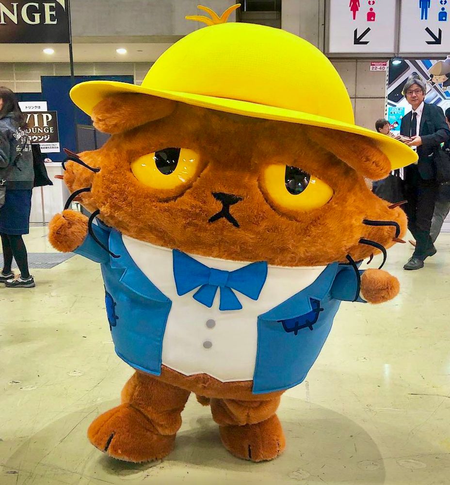 A drowsy-looking brown cat mascot wears a blue jacket and bow tie and a yellow hat.