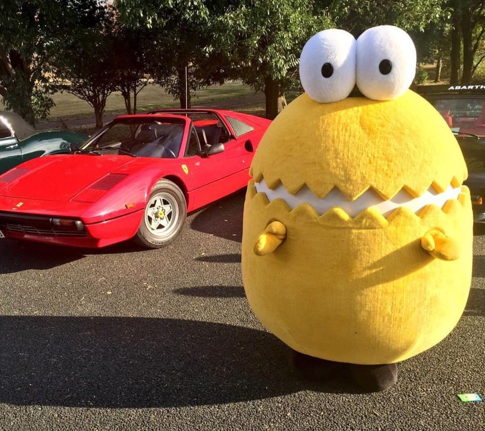 A yellow cracked egg mascot stands in front of a red sports car.