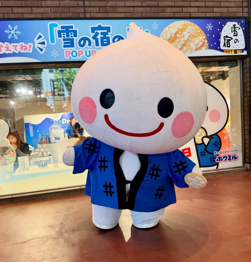 A smiling white mascot in a blue happi coat stands in front of a shop.