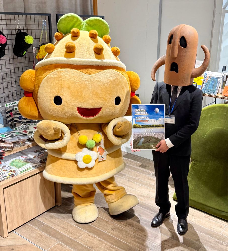 A smiling orange/brown mascot version of a haniwa figure wearing flower accessories stands behind a figure in a business suit, who has a haniwa head.