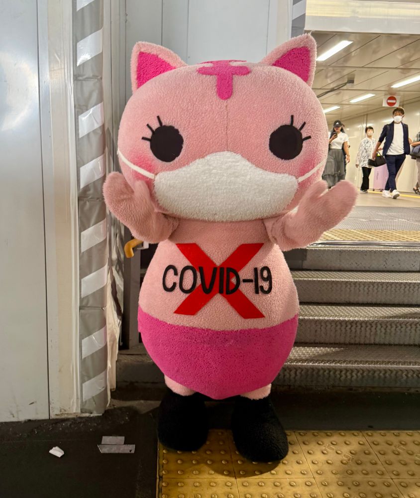 A pink cat mascot with a white face stands and waves in front of an urban train station.