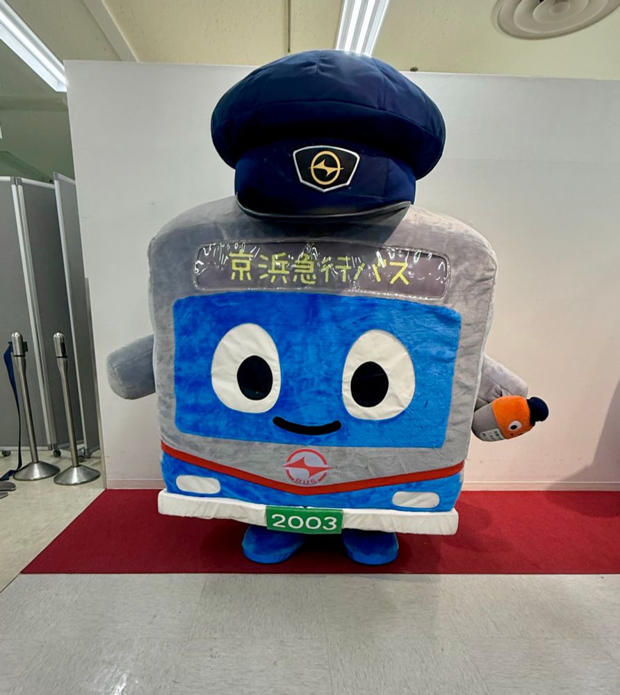 A smiling bus mascot in a blue cap stands on a red carpet.