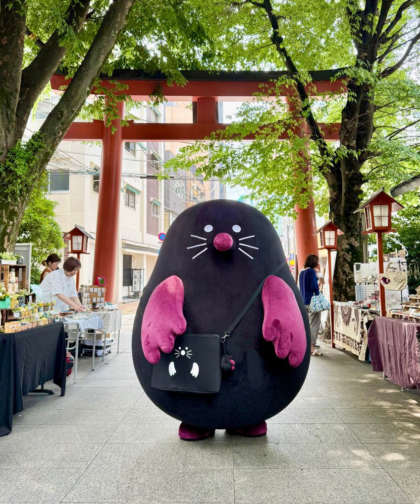 A black and purple mole mascot stands in front of a big red torii gate.