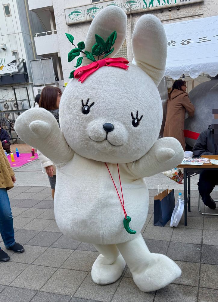 A white hare mascot with leaves around one ear and a bead around its neck, stands in the street.