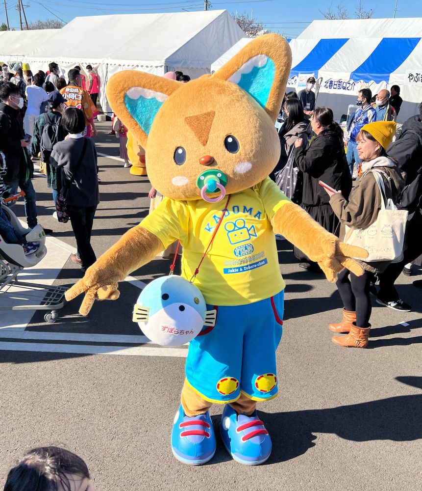 A fennec fox mascot with a pacifier, carrying a blue puffer fish like a handbag, stands among a crowd.