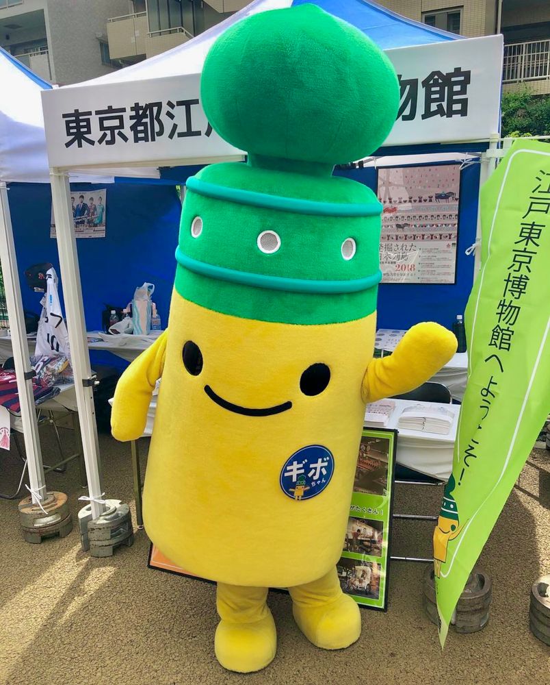 A yellow and green cylindrical mascot poses in front of its promotional booth.