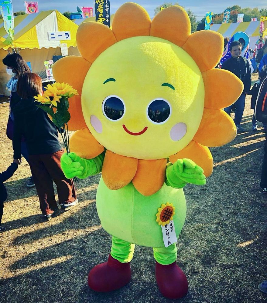 A smiling sunflower mascot holds a sunflower 