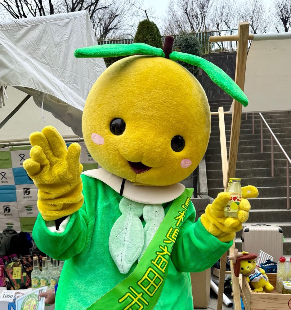 A mascot with a yellow fruit head and green clothes holds a bottle of juice in front of a staircase.