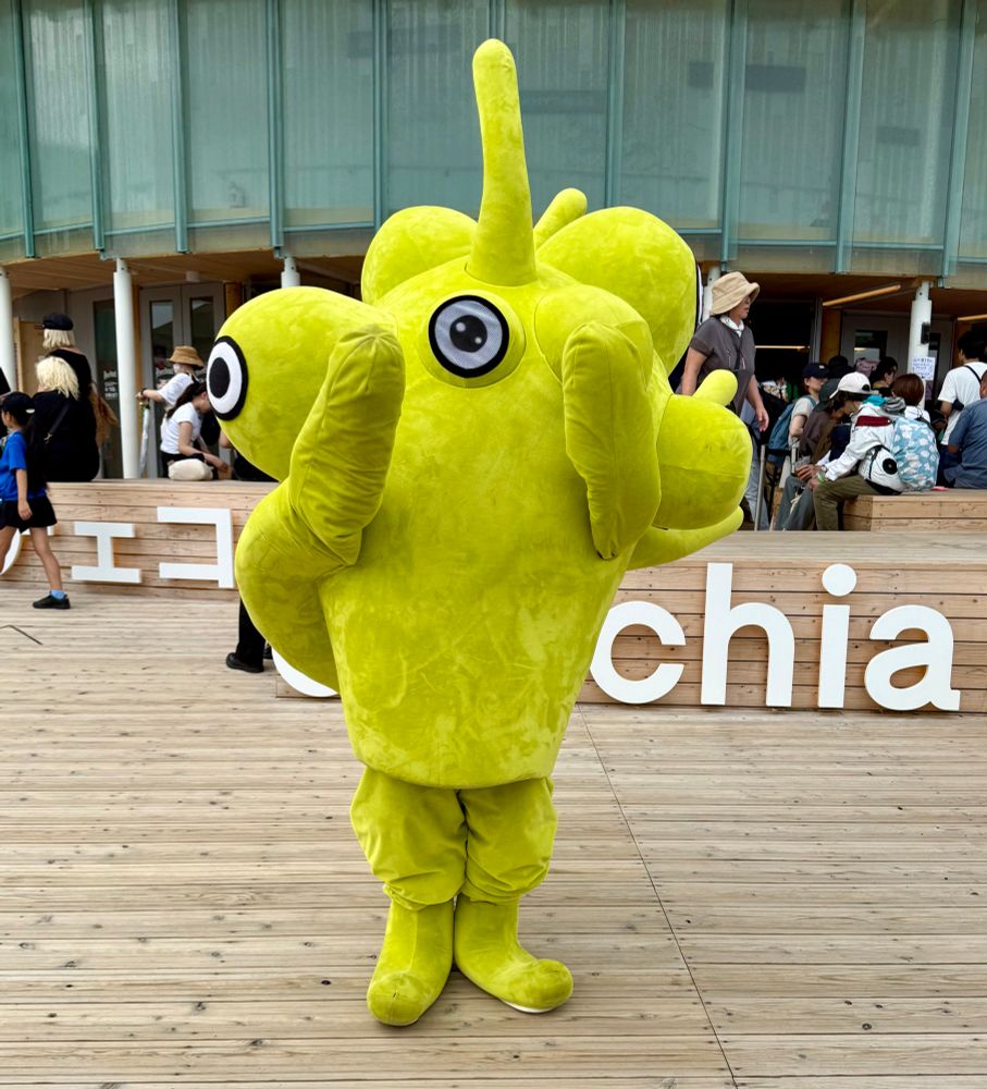 A yellow mascot with oddly-placed eyes and a protuberance on the top of its head stands on a wooden deck.